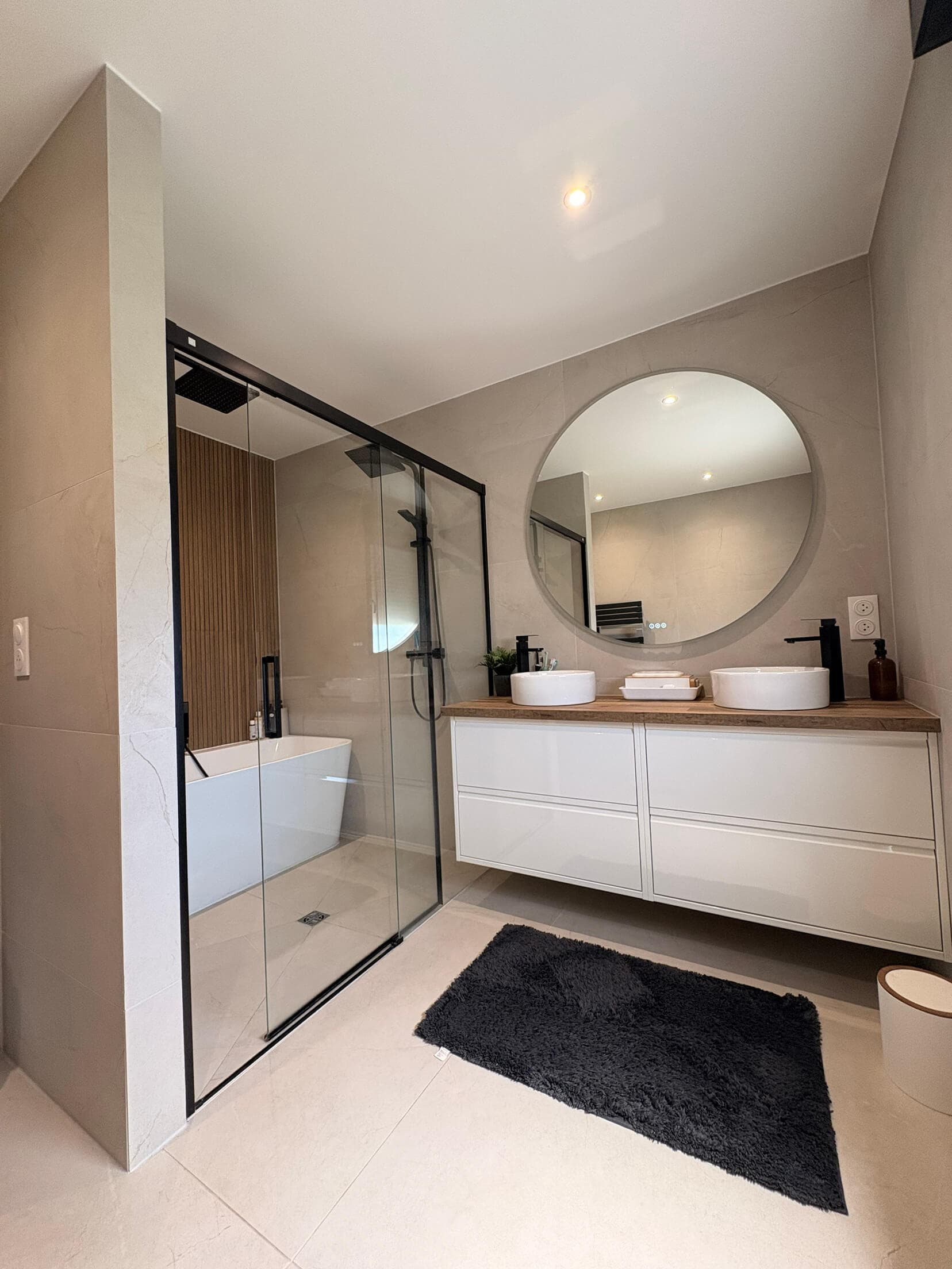 La salle de bain finie : douche vitrée, double vasque et miroir rond rétroéclairé